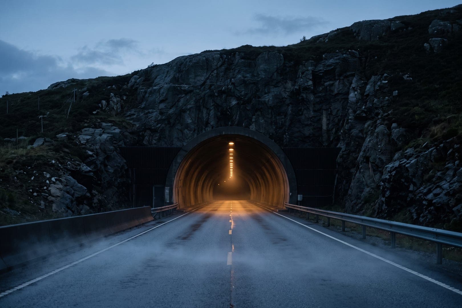 Boca de un túnel de carretera al anochecer, vista de frente a distancia, con una hilera de luces perdiéndose dentro y niebla baja sobre el asfalto.